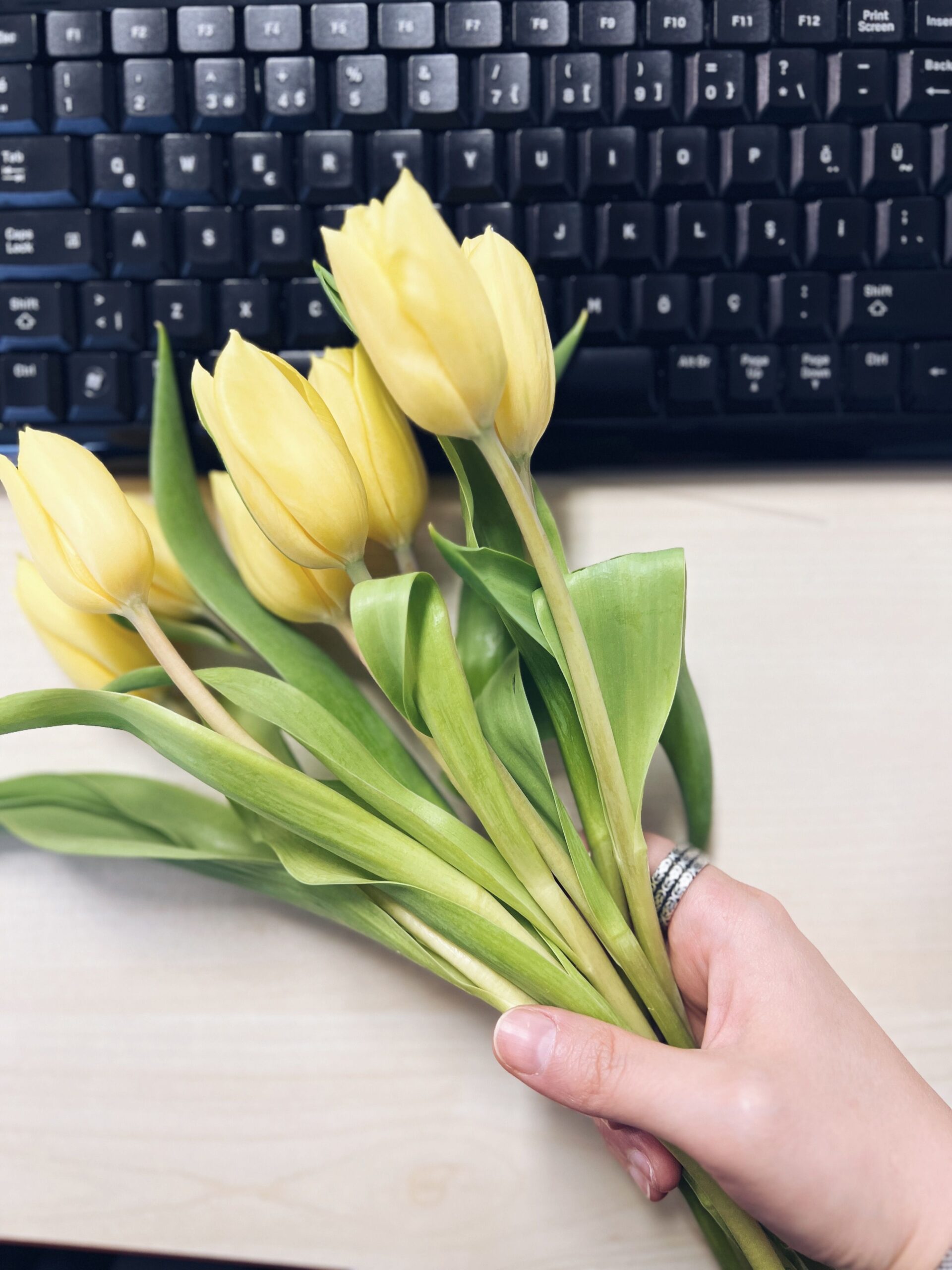 A hand holding yellow tulips above a keyboard, blending nature with technology in a workspace.